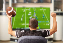 How to Get NFL Sunday Ticket in 2023 Man holding a football and wearing a football jersey cheers while watching a football game on TV