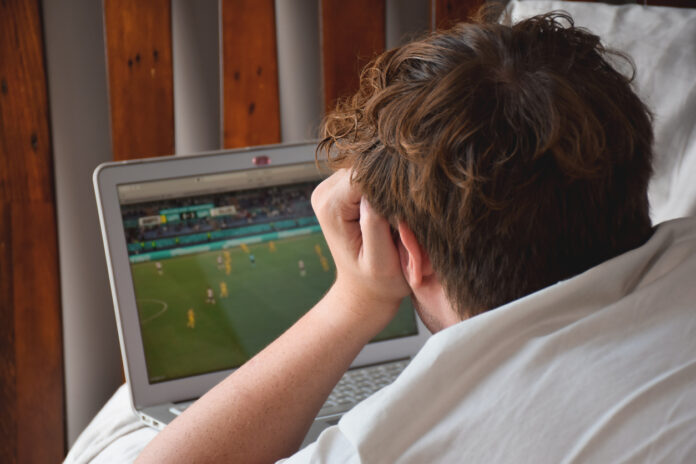 A young man laying on his bed watching an international soccer game on his laptop computer.