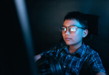 How to Teach Your Kids About Online Piracy and Its Risks Young kid on the computer playing games, watching entertainment or browsing the online digital internet, wearing glasses and dark background theme.