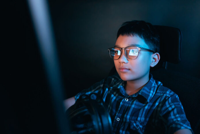 Kids’ Safety Young kid on the computer playing games, watching entertainment or browsing the online digital internet, wearing glasses and dark background theme.