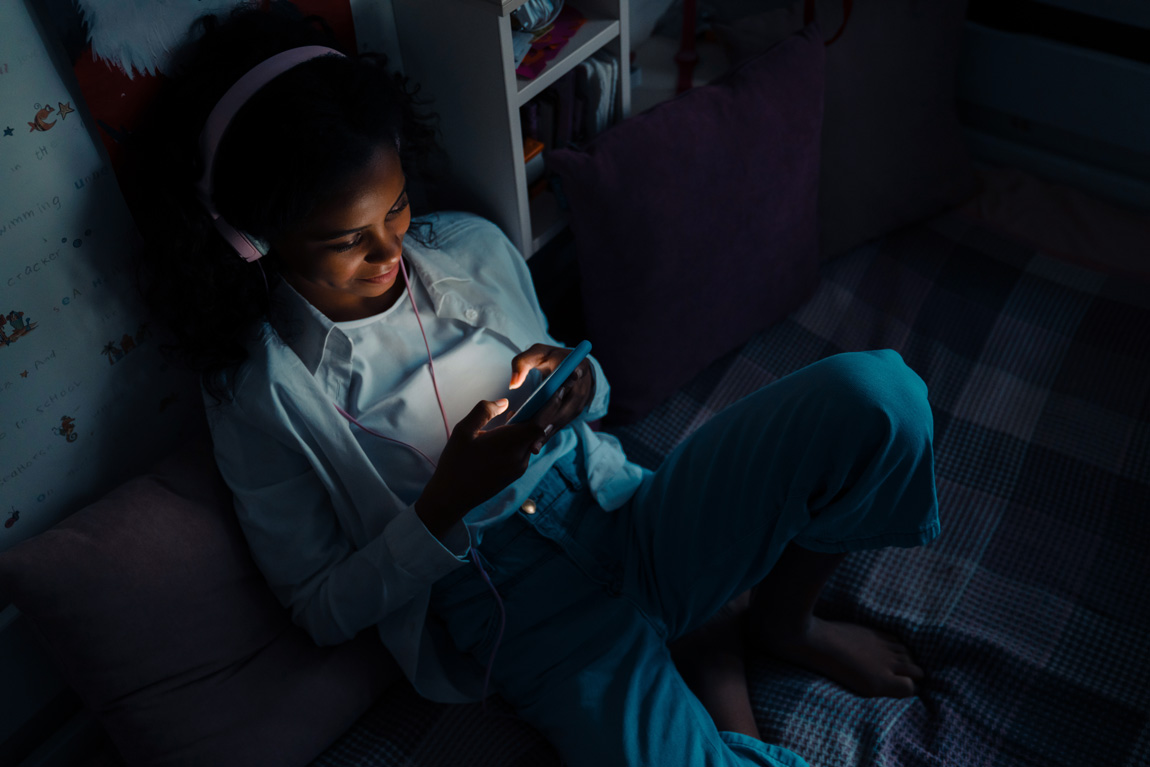 WhyIS_SafeStreaming Smiling teen girl sitting on a bed with phone texting or using mobile apps in her room at home