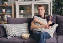 Is It Illegal To Stream Movies and TV Shows Online? What You Need To Know A man sitting on a sofa holding a pillow while watching TV, with a bowl of popcorn beside him.