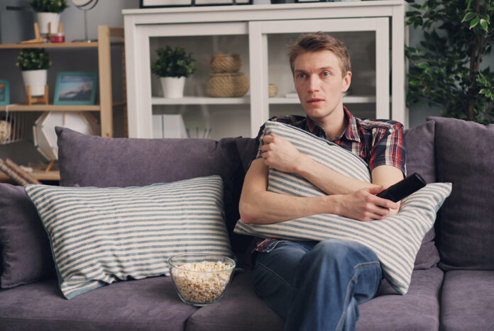 IsItIllegal A man sitting on a sofa holding a pillow while watching TV, with a bowl of popcorn beside him.