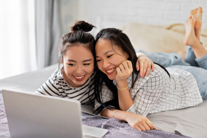 Two smiling women enjoying time together using a laptop in a cozy bedroom setting.